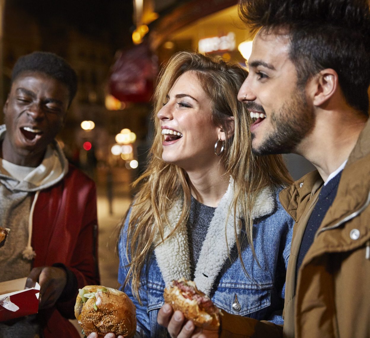 Laughing friends holding burgers at night. Young men and women are enjoying in city. They are wearing casuals.